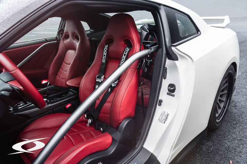 Close-up of a Nissan GT-R interior featuring red racing seats, Sparco harnesses, and a roll bar.