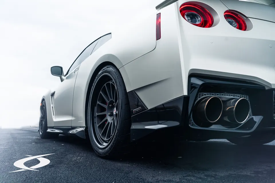 White Nissan GT-R with custom black multi-spoke wheels and large quad aftermarket exhaust tips, low angle rear view.
