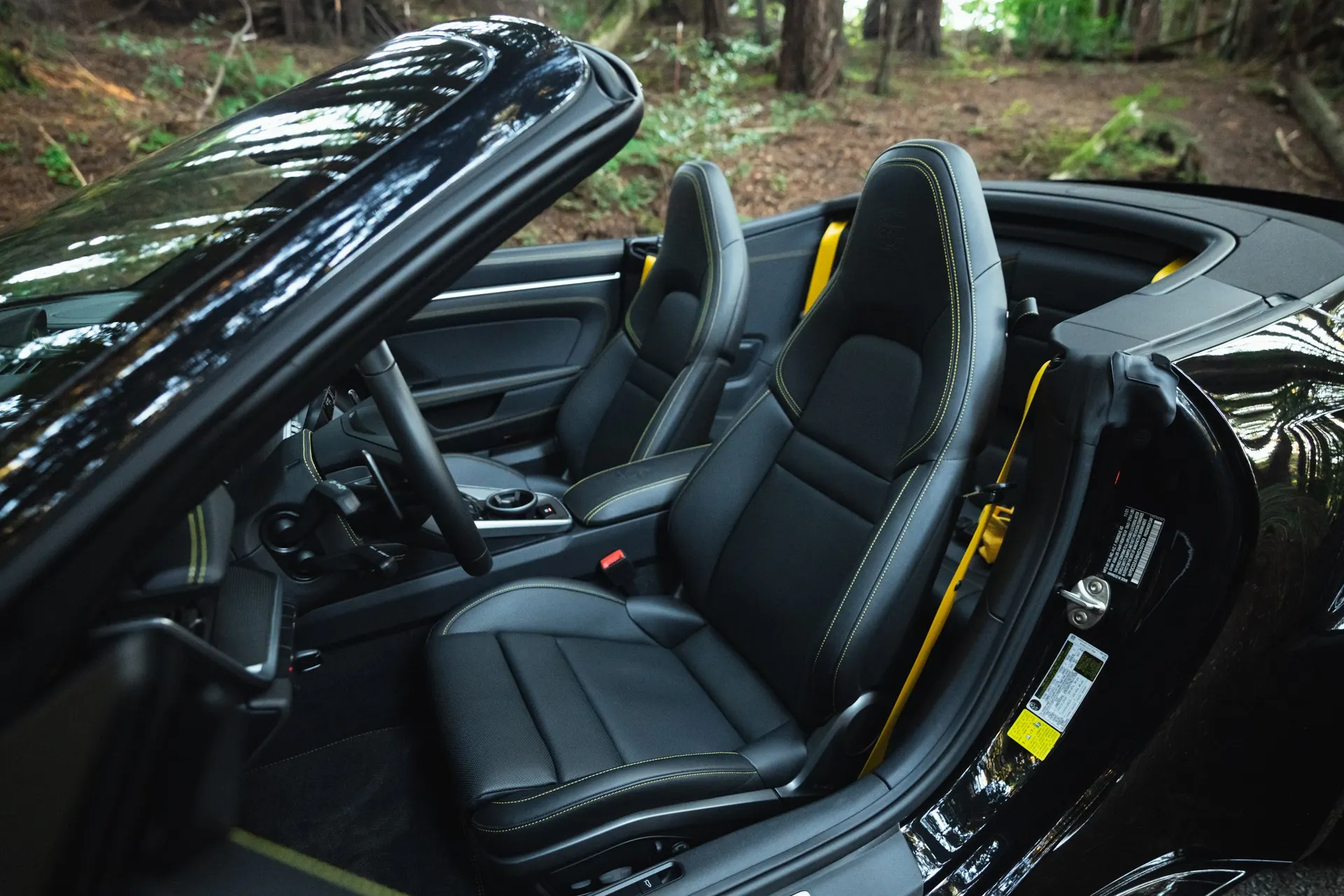 Interior view of a black Porsche convertible with black leather seats, yellow stitching, and yellow seatbelts, parked outdoors.