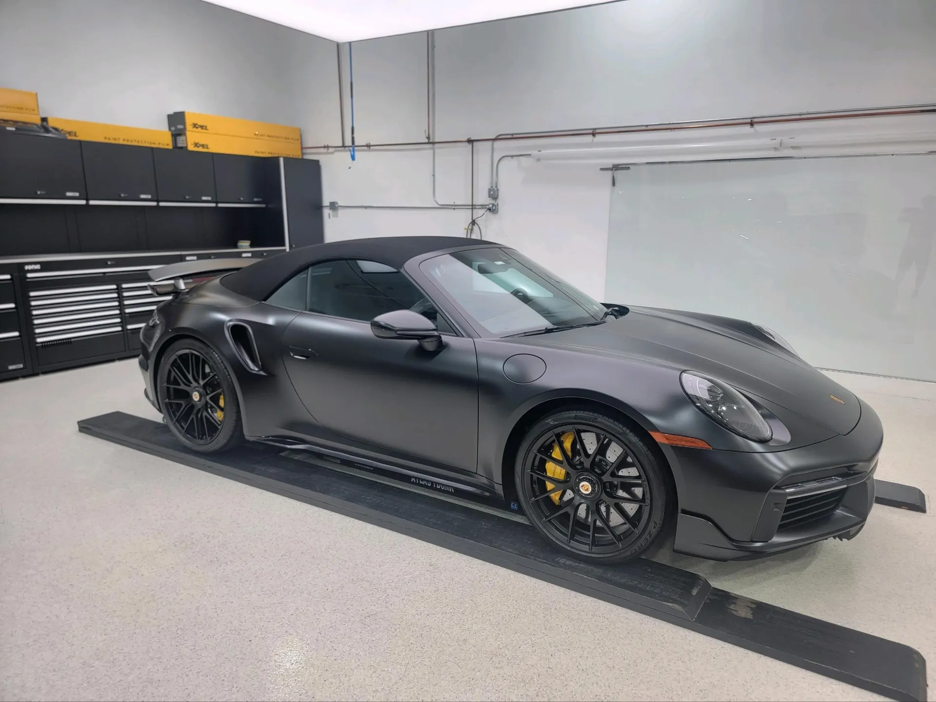 A matte black Porsche 911 Turbo S convertible with yellow brake calipers parked on ramps in a clean shop.