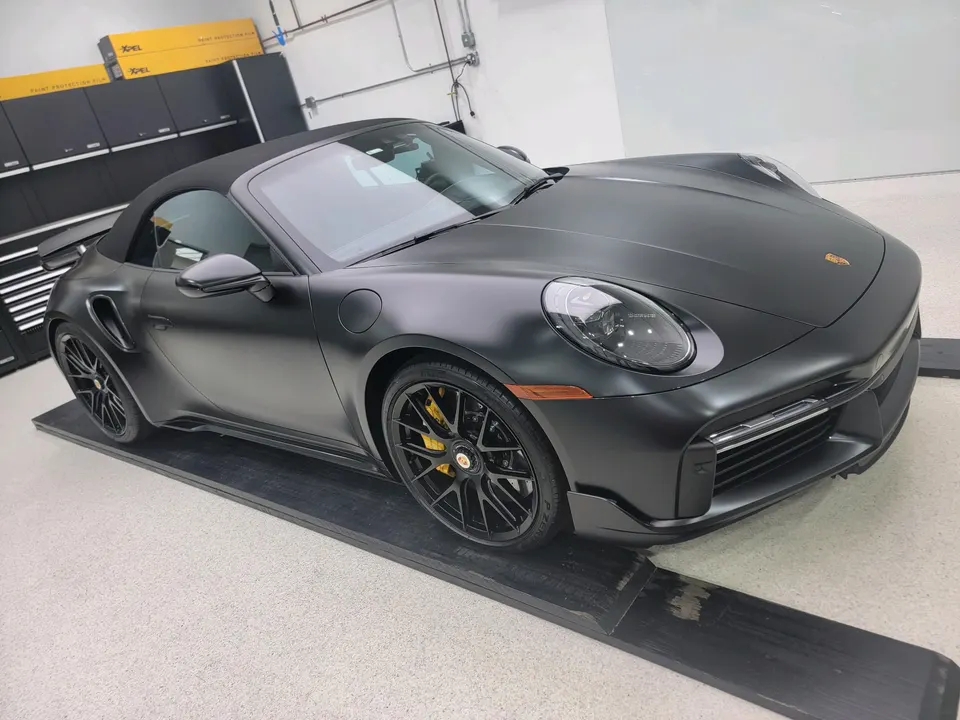 Matte black Porsche 911 Turbo S convertible on ramps in a shop with XPEL paint protection film boxes in the background.