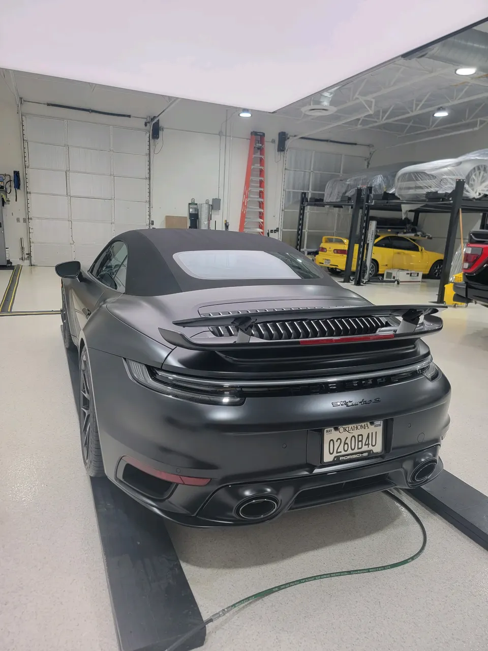 Rear view of a matte black Porsche 911 Turbo S convertible in a performance tuning shop with other cars on lifts.