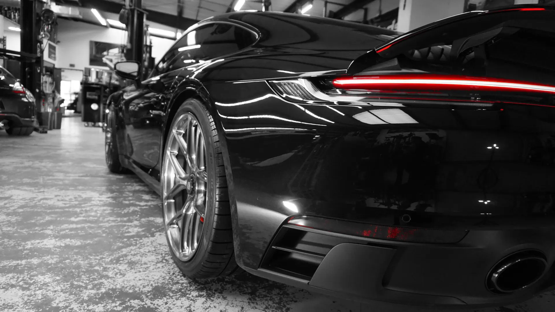 Rear view of a black Porsche 911 parked in a well-lit performance tuning shop, showcasing its sleek design and wheel.