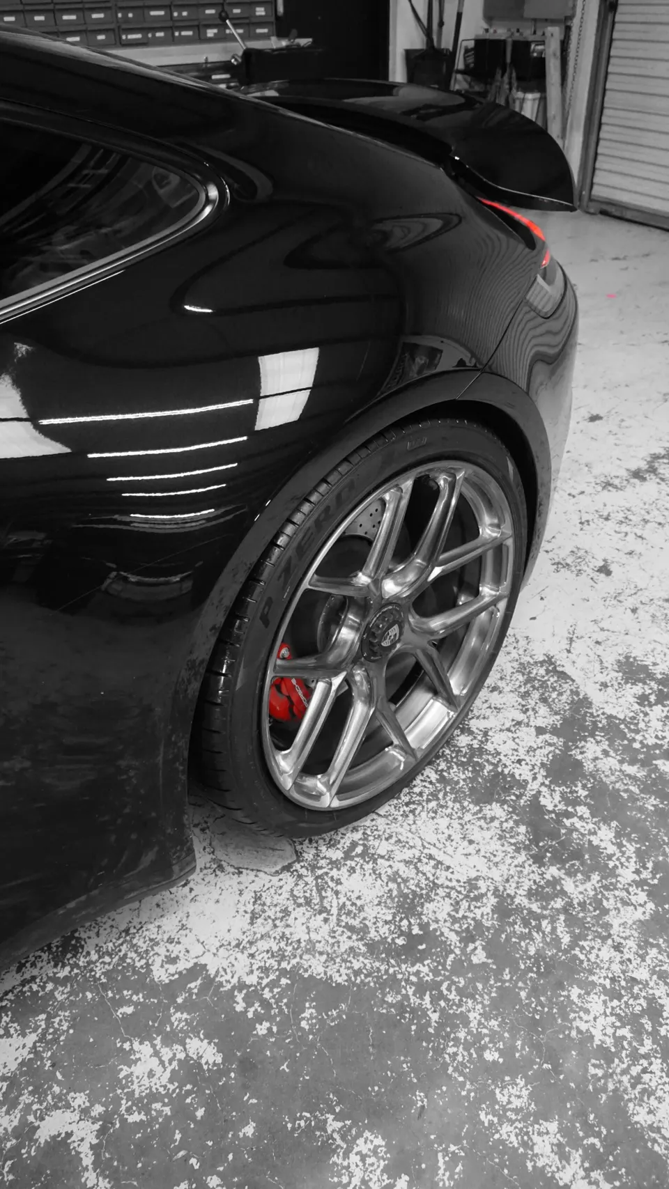 Black Porsche rear quarter panel with custom multi-spoke wheels, red brake calipers, and Pirelli P Zero tires on a shop floor.