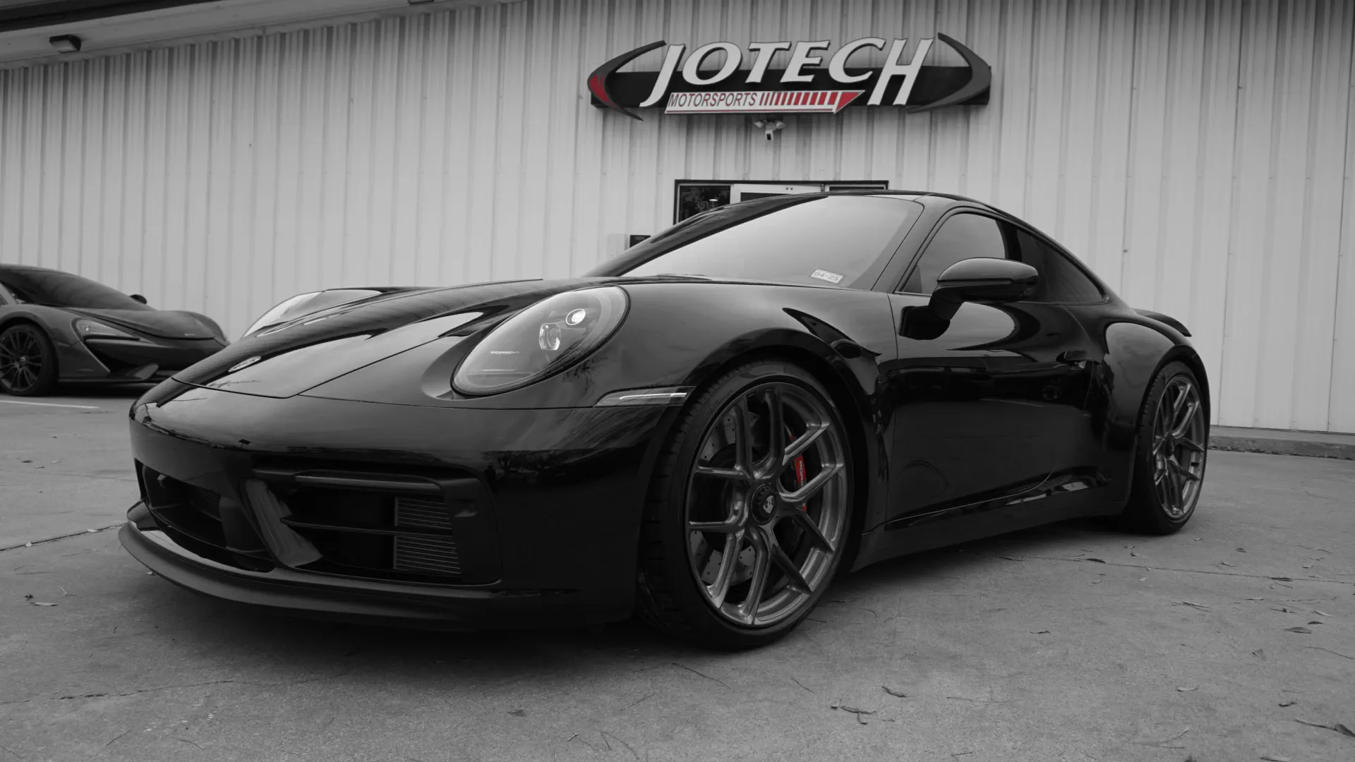 Black Porsche 911 parked outside Jotech Motorsports shop with a McLaren in the background.