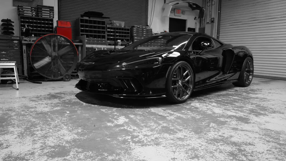 A sleek black McLaren supercar parked inside a garage with an industrial fan and storage shelves in the background.