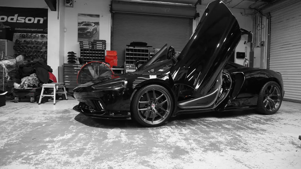 Black McLaren supercar with butterfly doors open, parked inside a performance tuning shop with tools and equipment.