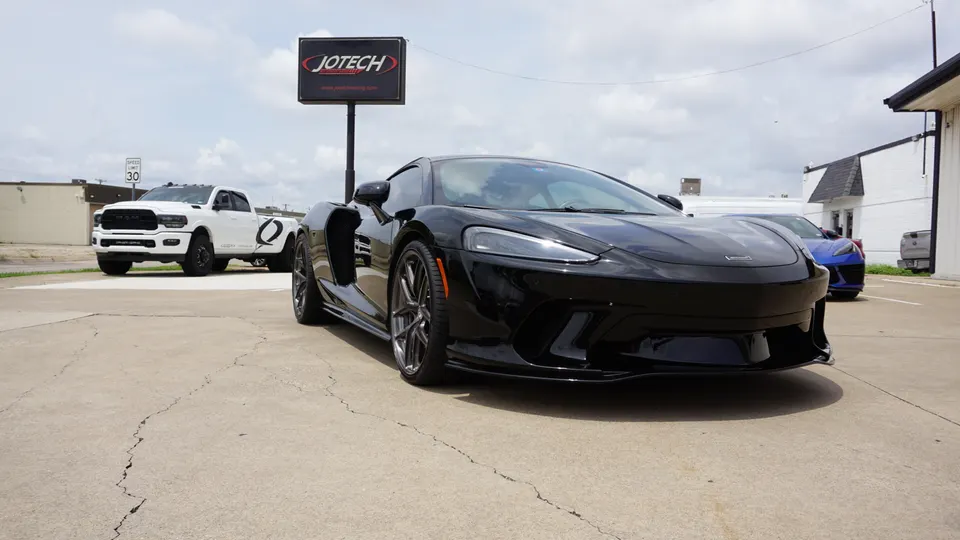 A black McLaren supercar parked outside Jotech Motorsports, with a white Ram truck in the background.