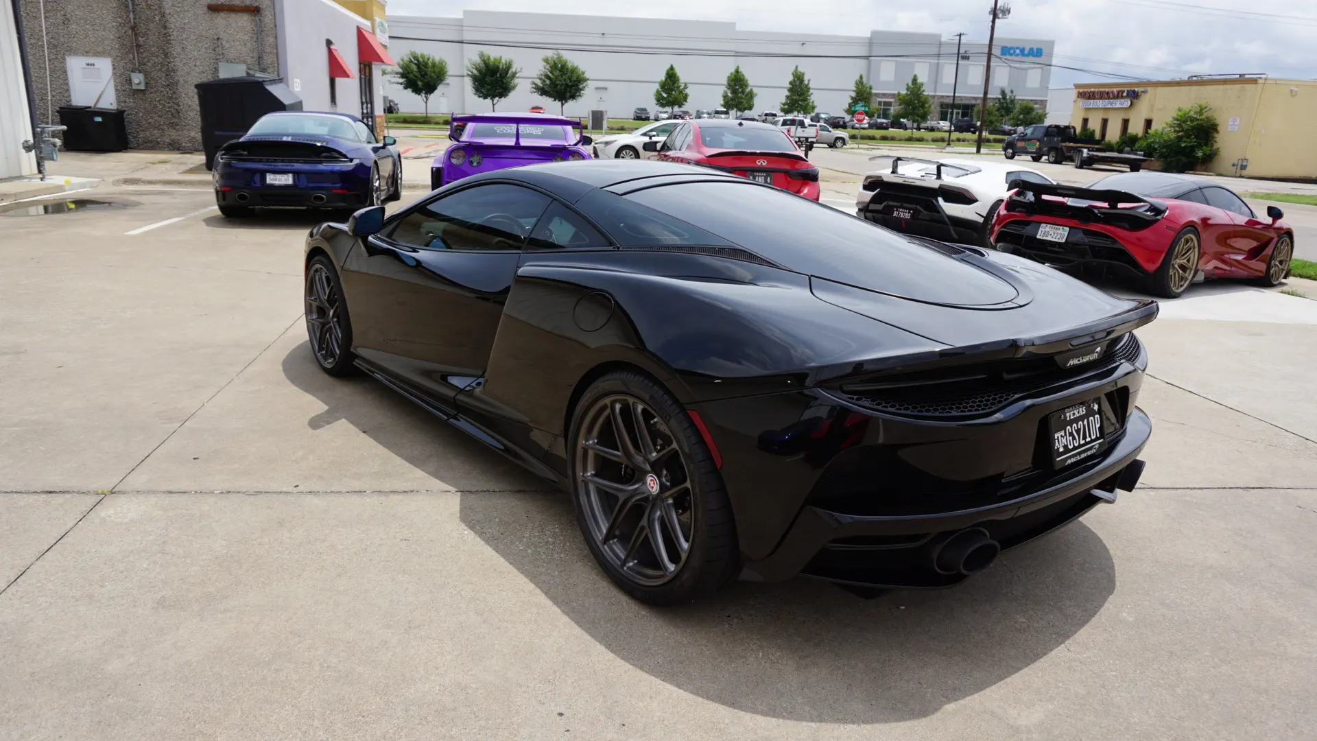 A black McLaren parked in a lot with other supercars including a red McLaren, purple GT-R, blue Porsche, and white Lamborghini.