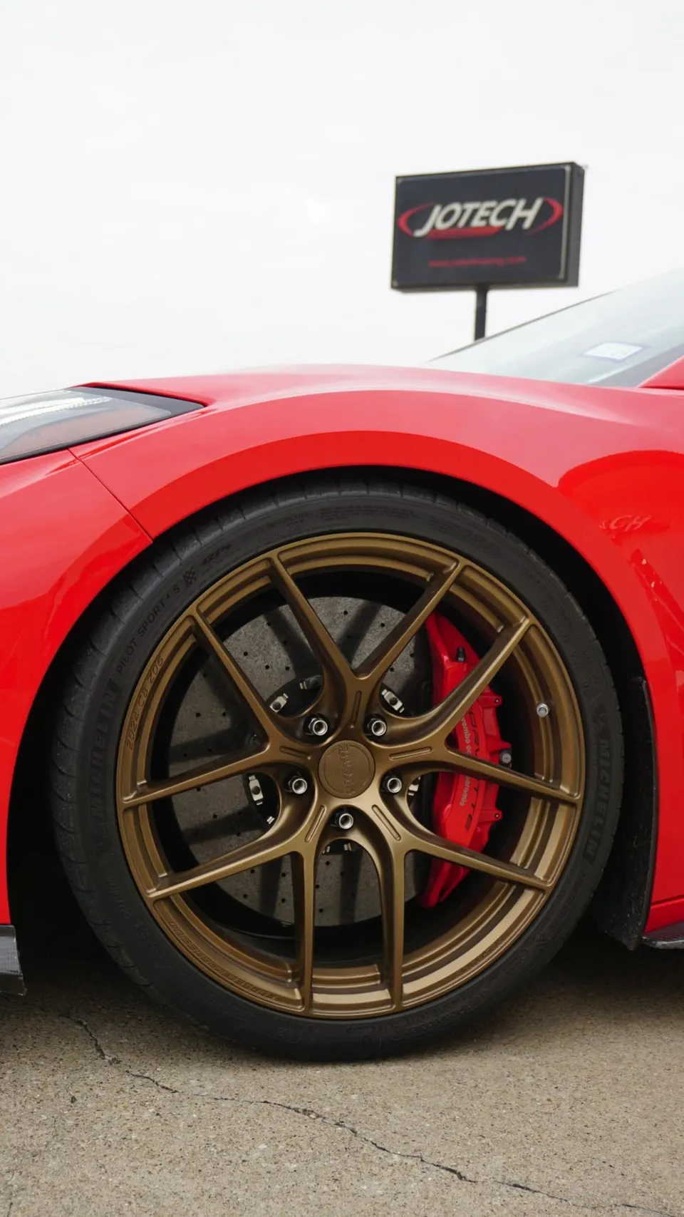 A close-up of a red car's bronze multi-spoke wheel with red Brembo brake calipers and Michelin tires, with a Jotech sign in the background.