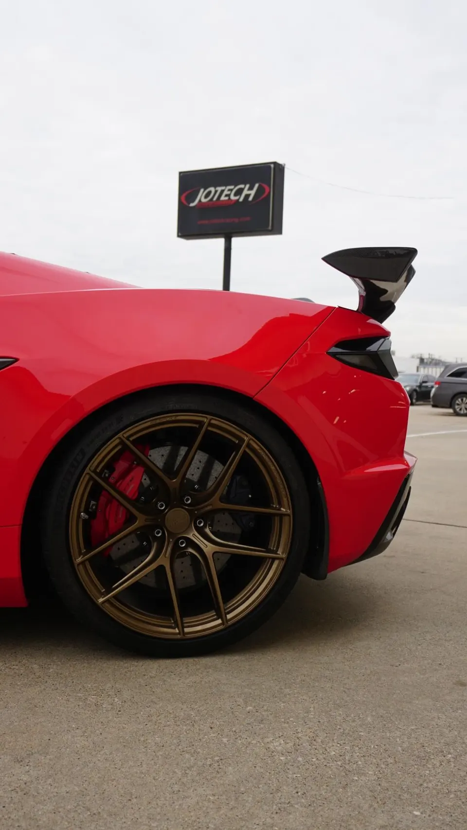 Red C8 Corvette with bronze aftermarket wheels, red brake calipers, and a carbon fiber rear wing parked outside Jotech Motorsports.