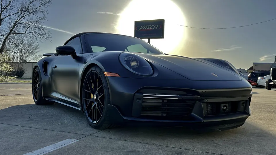 A matte black Porsche 911 parked outdoors with a Jotech sign in the background, showcasing its sleek design.