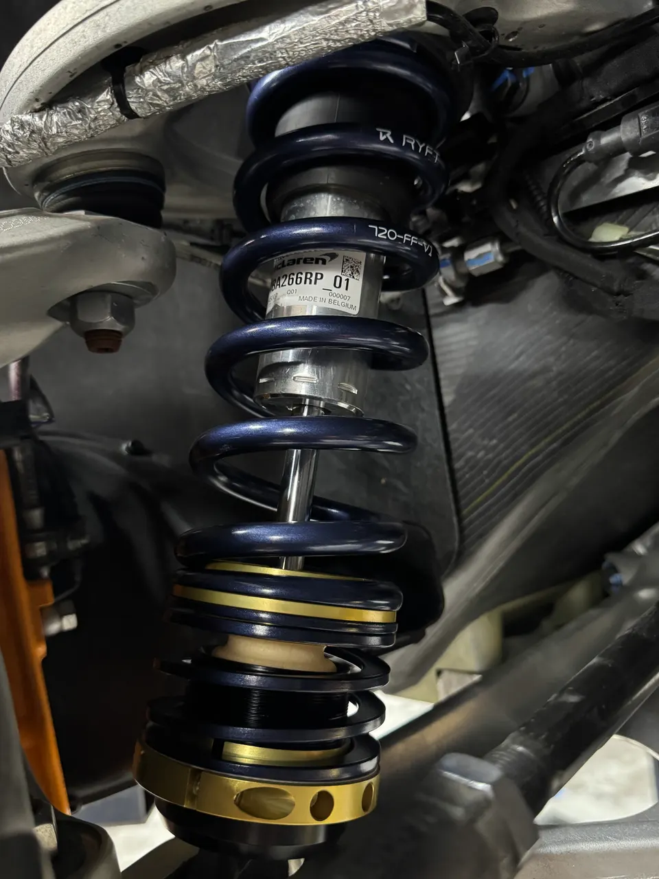 Close-up of a McLaren coilover suspension with blue springs and gold adjusters, showing part number label.