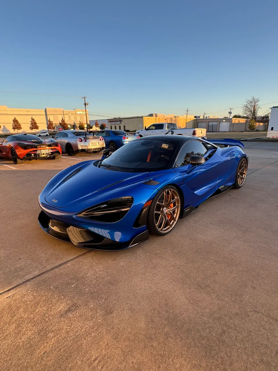 Blue McLaren 720S with bronze aftermarket wheels parked outdoors at golden hour, with other supercars in the background.