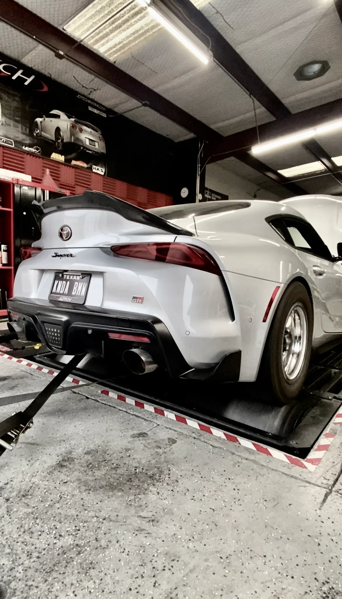 A silver Toyota Supra MK5 strapped to a dyno at Jotech Motorsports, showing its rear with a custom license plate and exhaust.