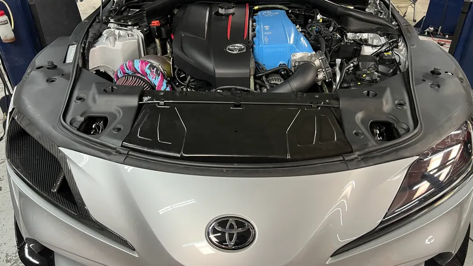 Front view of a silver Toyota Supra with its hood open, showcasing a modified engine bay with a blue intake manifold, aftermarket turbocharger, and colorful air