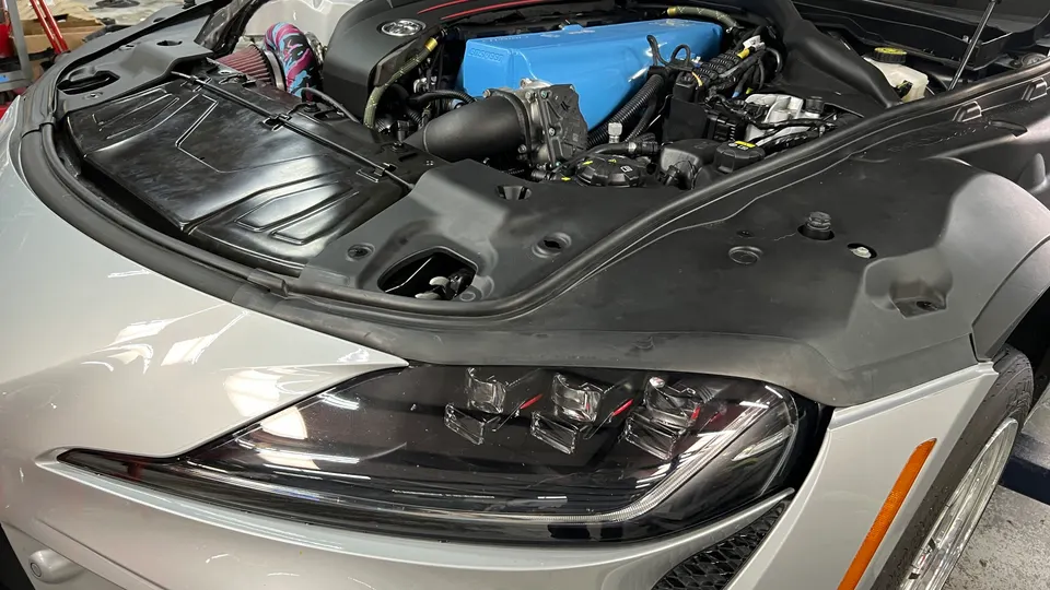A silver Toyota Supra with its hood open, showcasing a blue aftermarket intake manifold and an upgraded air intake system in the engine bay.