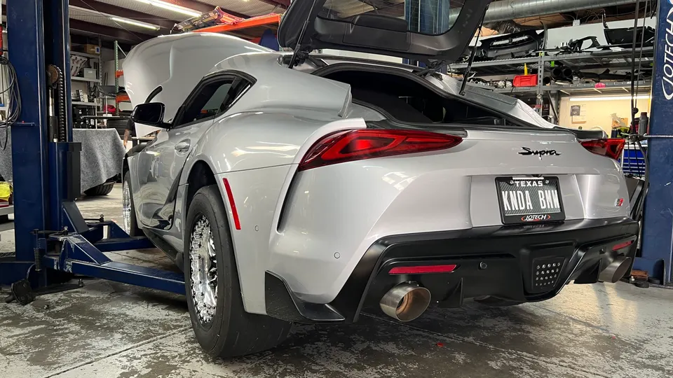 A silver Toyota Supra MK5 on a lift in a performance shop, featuring drag wheels and an aftermarket exhaust.