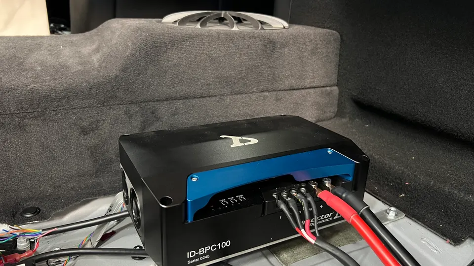 Close-up of an ID-BPC100 audio amplifier installed in the trunk of a car with visible wiring connections.