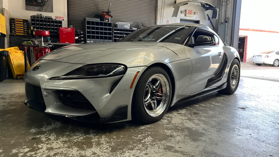 Silver Toyota Supra with aftermarket drag wheels parked inside a performance tuning shop.