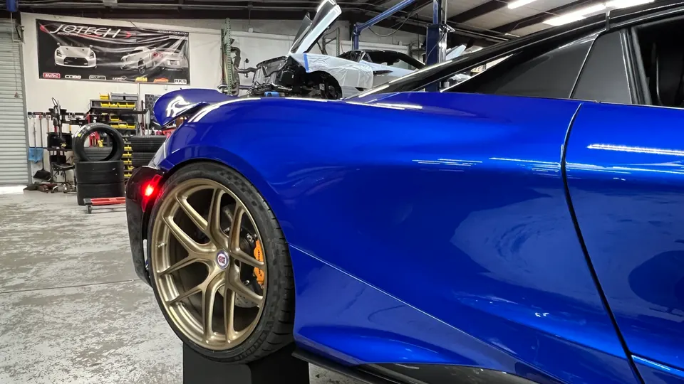 A blue McLaren supercar with gold wheels and orange brake calipers in a Jotech Motorsports performance shop.