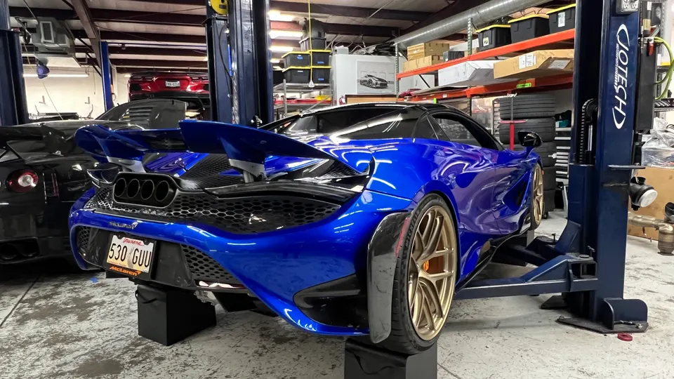 Blue McLaren 720S with a large active rear wing and quad exhaust tips, on stands in a performance tuning shop.