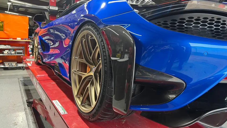 A blue McLaren supercar with gold aftermarket wheels and carbon fiber aero on a red car lift in a performance tuning shop.