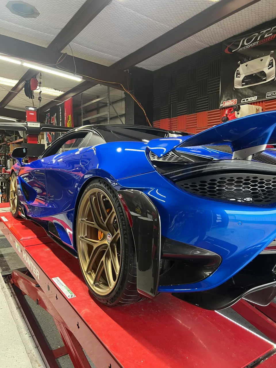Blue McLaren 720S on a red shop lift, featuring gold aftermarket wheels and carbon fiber aerodynamic parts.