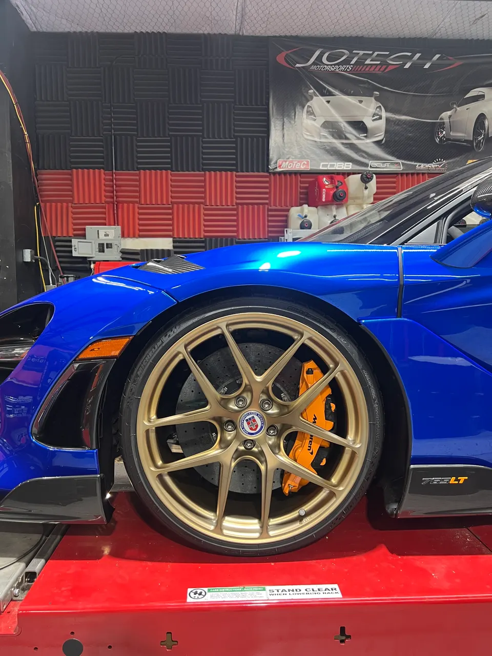 A blue McLaren 765LT with gold HRE wheels and orange brake calipers on a red dyno at Jotech Motorsports.