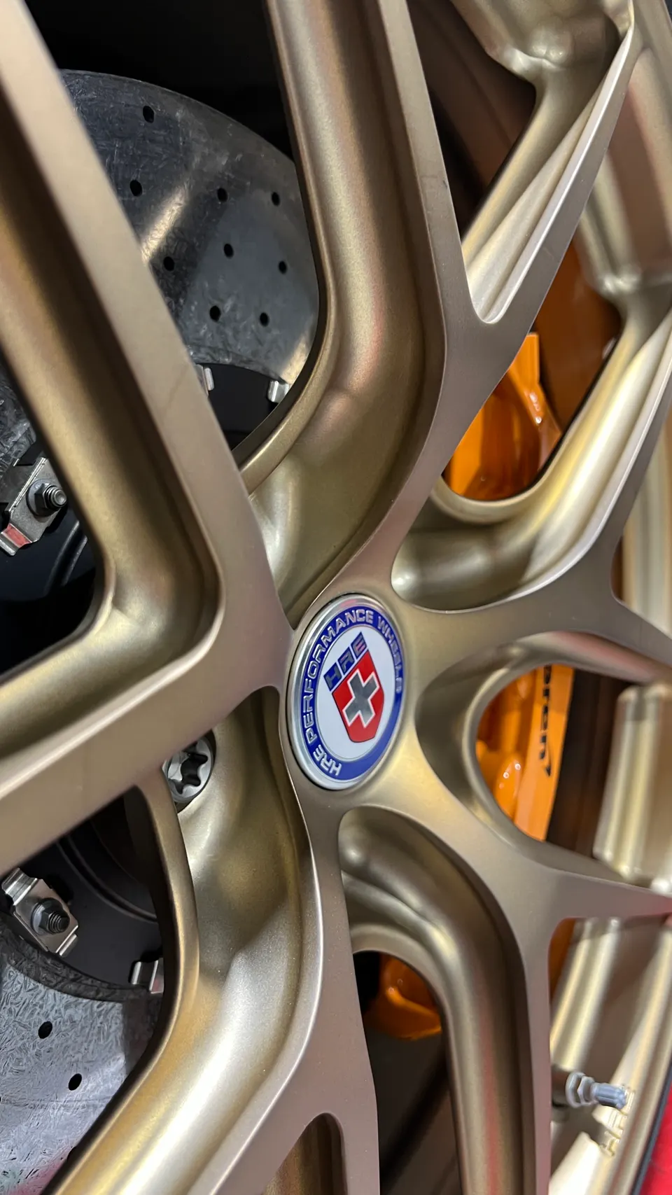 A close-up of a gold HRE performance wheel with orange McLaren brake calipers and carbon ceramic rotors.