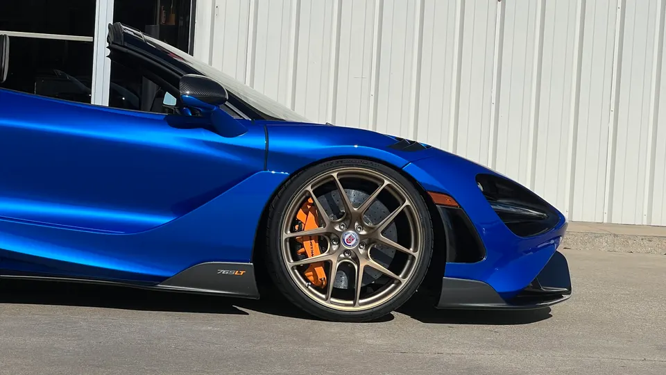 A blue McLaren 765LT supercar with bronze HRE wheels and orange brake calipers parked against a white wall.