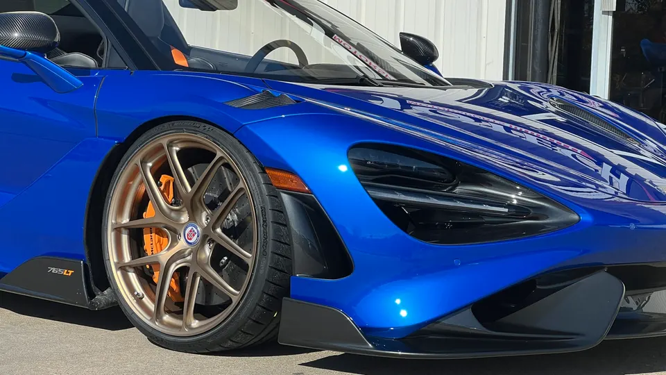Blue McLaren 765LT with bronze HRE wheels and orange brake calipers, showcasing the front fender and headlight.