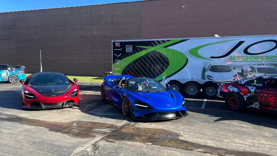 Two McLaren 720S supercars, one red and one blue, parked in front of a Jotech Motorsports trailer with other custom cars.