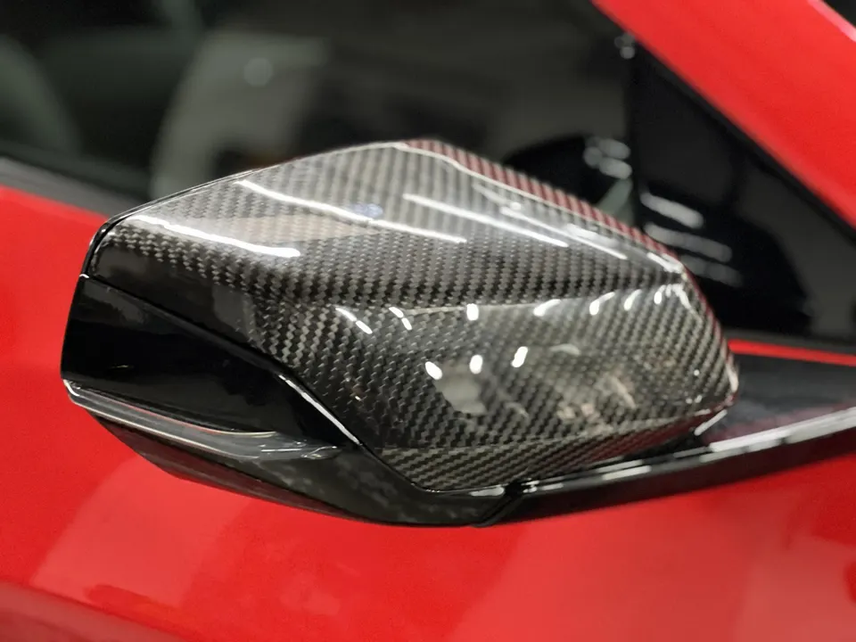 Close-up of a carbon fiber side mirror on a red sports car, showcasing the weave pattern and glossy finish.