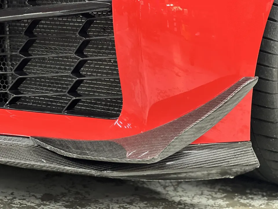 Close-up of a red car's front bumper with a carbon fiber splitter and canards, showcasing aerodynamic modifications.