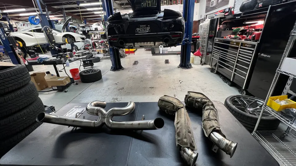 A black McLaren on a lift in a performance tuning shop, with a new stainless steel exhaust system and old exhaust parts on a table.