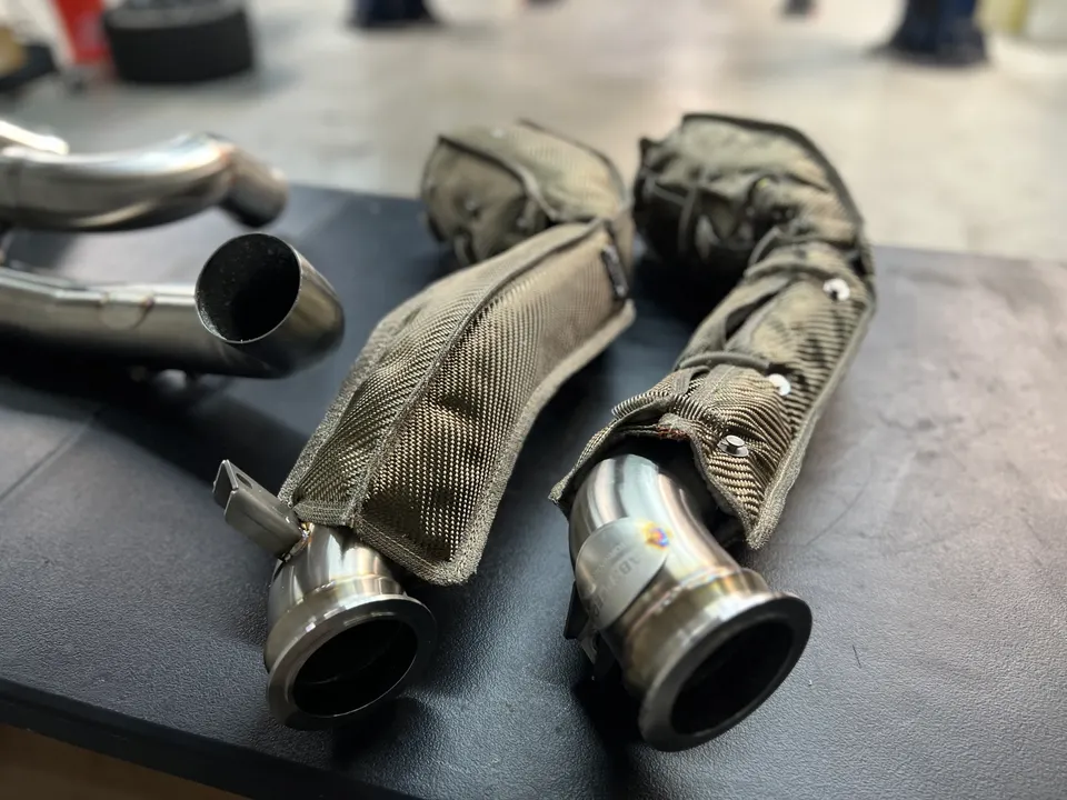 Close-up of performance exhaust downpipes with heat shields on a workbench in a tuning shop.