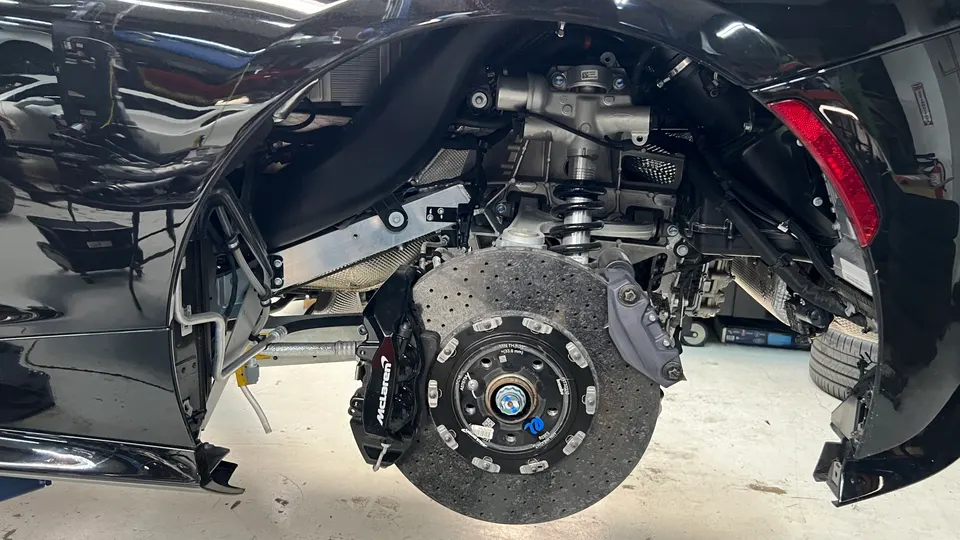 A McLaren supercar in a shop with the front wheel removed, showcasing the carbon ceramic brake rotor, caliper, and suspension components.