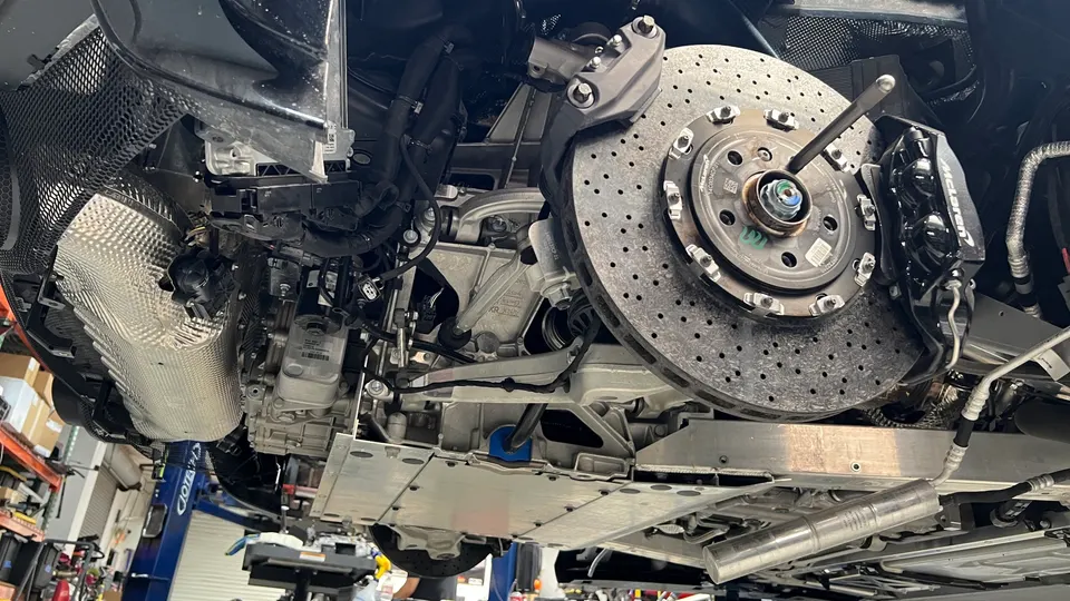 Underside view of a McLaren on a lift in a shop, showcasing the large brake rotor, caliper, and suspension components.