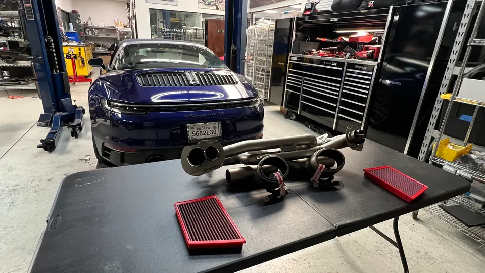 Blue Porsche Carrera GTS in a performance shop with an aftermarket exhaust system and air filters on a table.