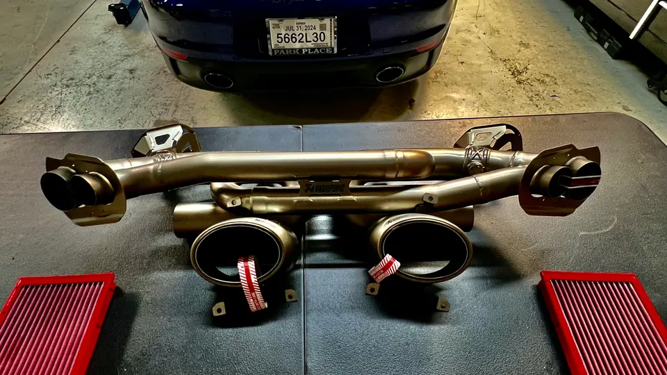 Akrapovic exhaust system and performance air filters laid out in a shop with a Porsche in the background.