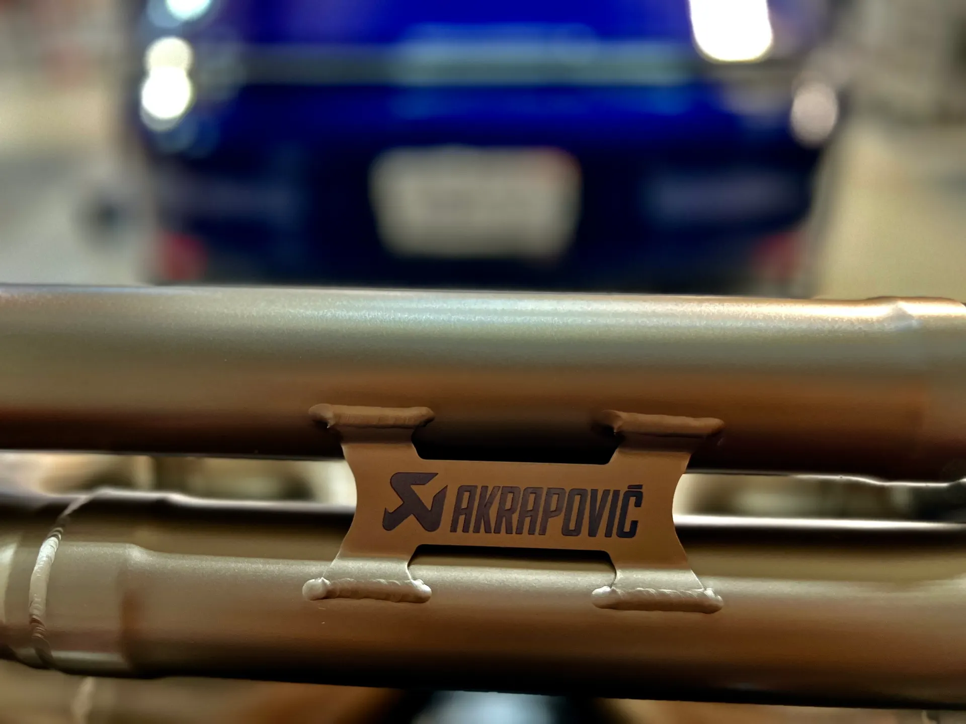 Close-up of an Akrapovic exhaust system with a blurred car in the background, highlighting the brand logo and welds.