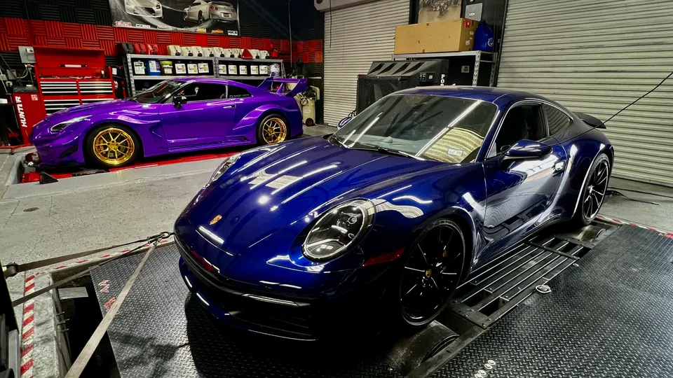 A blue Porsche and a purple Nissan GT-R strapped to a dynamometer in a performance tuning shop.