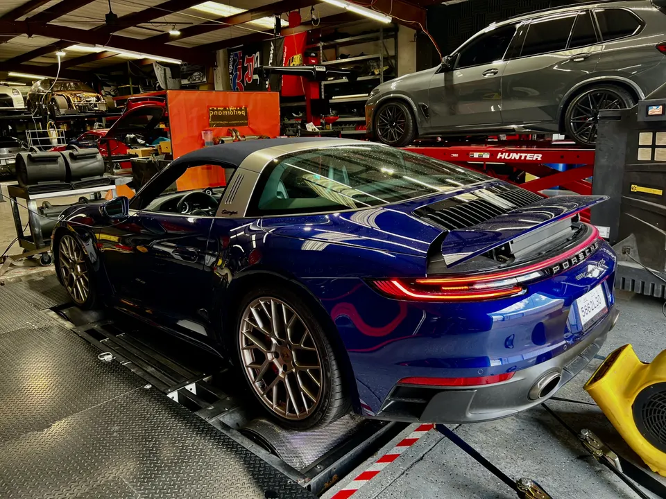 Blue Porsche 911 Targa 4S on a dyno at a performance tuning shop, with a grey SUV on a lift in the background.