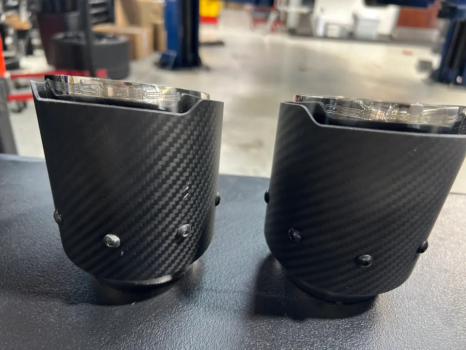 Two carbon fiber exhaust tips with Porsche branding on a workbench in a performance tuning shop.