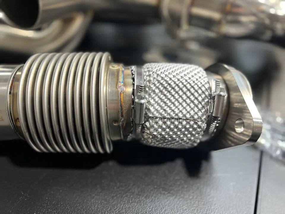 A close-up of a stainless steel automotive exhaust flex pipe with a mesh heat shield.