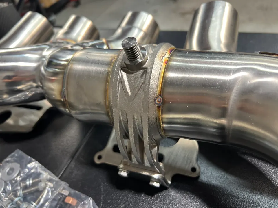 Close-up of a stainless steel performance exhaust component with visible welds and a clamp, likely a downpipe or midpipe.