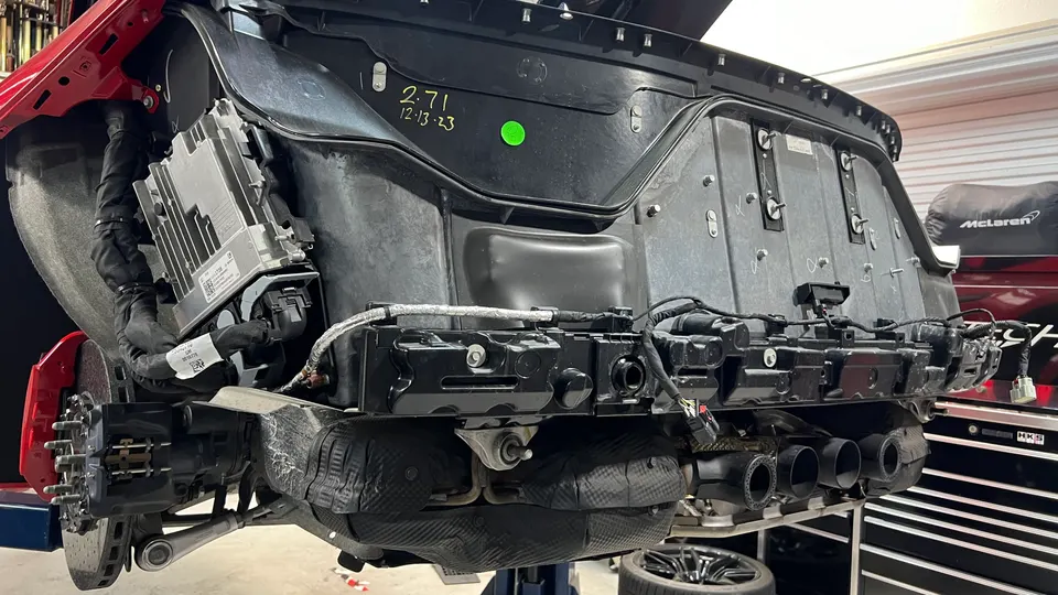 Rear view of a McLaren on a lift in a shop, with the bumper removed, showing the exhaust system, brakes, and electronic control unit.