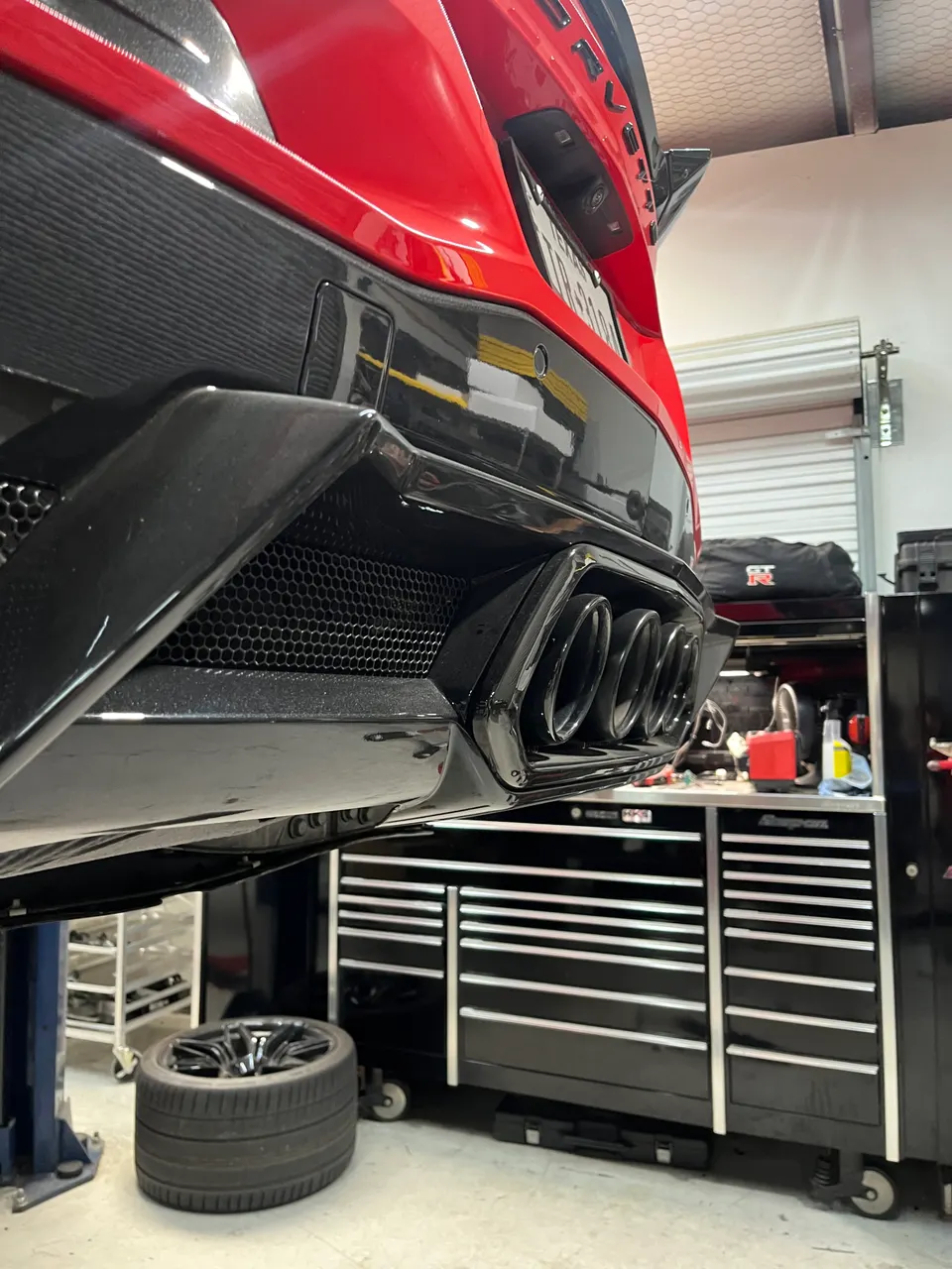A red Corvette C8 on a lift in a shop, showcasing its quad exhaust tips and carbon fiber rear diffuser.