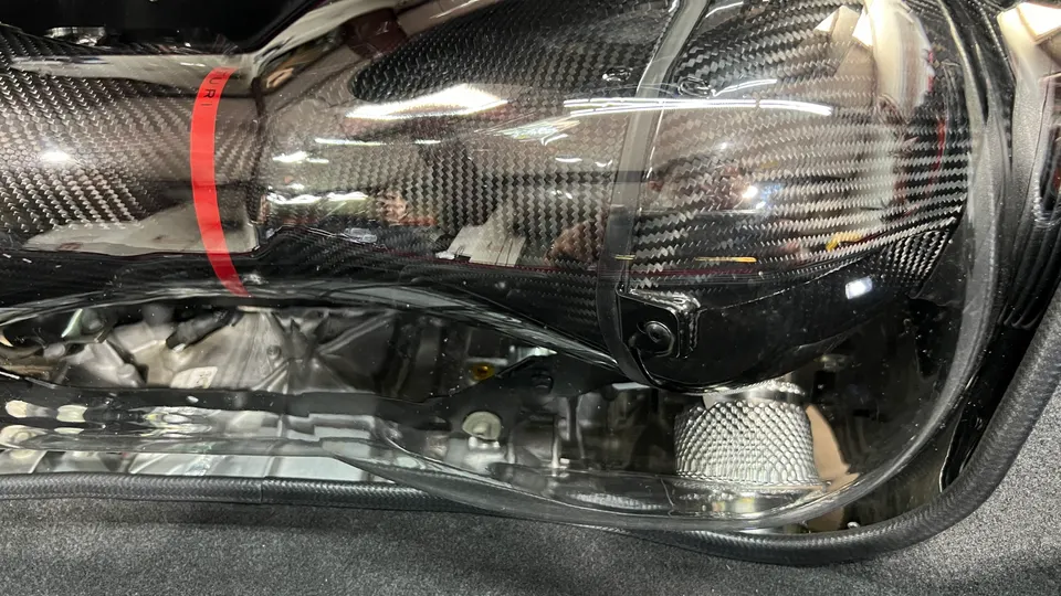 Close-up of a carbon fiber air intake system with a red stripe, visible through a clear cover in an engine bay.